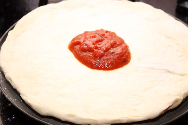 После второго подъема можно приступать к приготовлению пиццы. Тесто раскатать (или понемногу растянуть) до размера формы, в которой вы будете готовить. Толщина теста должна быть примерно 0,5-1 см. Это зависит от того, какую основу вы предпочитаете — потолще или потоньше.  На тесто выложить томатный соус для пиццы и равномерно распределить его по основе.