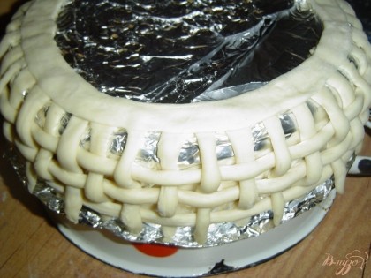 Пока торт находится под прессом займемся украшением. Прежде всего изготовим корзинку, для этого слоеное тесто нарезаем полосками, 4 длинных, а остальные нарезаем по высоте торта + 2 см. Берем миску диаметром на 2-3 см больше, чем диаметр торта, накрываем фольгой, сверху крепим короткие полоски, а между ними в шахматном порядке пропускаем длинные полоски и скрепляем концы. Одну длинную полоску немного раскатываем, с одной стороны немного смачиваем водой, накладываем на закрепленные короткие полоски и немного прижимаем.