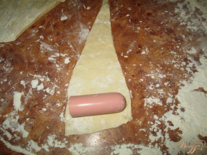 Затем, на посыпанной мукой поверхности, раскатываем тесто и вырезаем несколько треугольников, по количеству сосисок. На каждый треугольник из теста кладем  сосиску с широкой стороны.