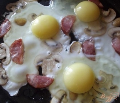 Когда грибы с колбаской запахнут, вбиваем три куриных яйца и сразу же соли, перчим по вкусу. Готовим яичницу до того момента, пока на яичном белке не пропадет слизь. Для ускорения процесса приготовления можно накрыть сковородку крышкой.