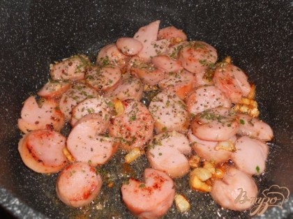 Рядом в сотейнике обжарить измельченный чеснок, добавить порезанные сосиськи и обжарить до золотистого цвета, добавить травки.