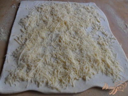 Слоёное тесто разморозить. Стол подпылить мукой, тесто раскатать (раскатывать в одном направлении).Сыр натереть на тёрке. Тесто посыпать сыром, сыр прижать рукой.