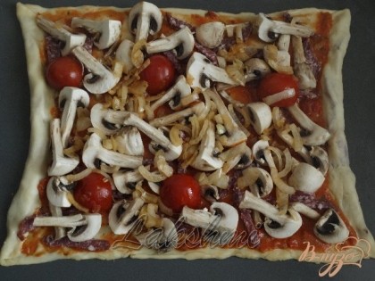 Основу пиццы смазываем приготовленным соусом. Выкладываем оставшиеся кусочки салями и шампиньоны. Отдельно на оливковом масле обжариваем лук до прозрачности, добавляем помидоры-черри. Выкладываем на тесто. Посыпаем итальянскими травами.