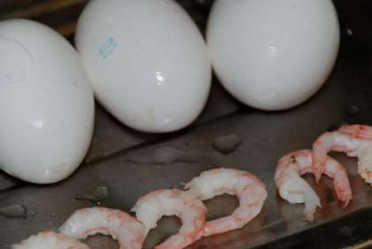 Чтобы сварить морепродукты нужно закипятить в кастрюле воду. Креветки опустить в подсоленную воду и держать их там  минуты 4. Затем почистить креветки и порезать на четыре части. Теперь необходимо сварить яйца вкрутую. Варить нужно минут 8 в соленой воде. Затем яйца остудить и почистить.