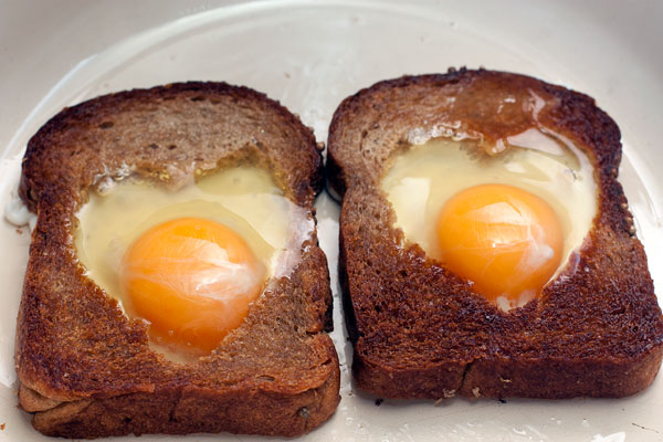 В хлебные окошки аккуратно разбейте яйца, стараясь не повредить желток. Жарьте на небольшом огне до нужной степени готовности.