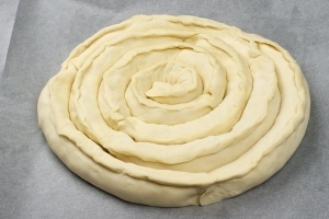 Каждую полоску закрутите по спирали, сформируйте круглый пирог. Противень застелите пергаментной бумагой, выложите на него пирог и смажьте верх взбитым яйцом. Сверху присыпьте кунжутом и поставьте в разогретую духовку до 200 градусов. Выпекайте пирог на протяжении 25 минут.