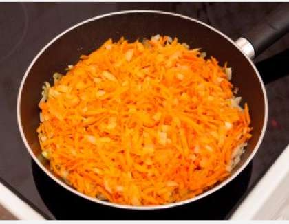 Разогрейте сковороду с высокими бортами. Положите на дно жир с тушенки. Влейте немного воды. Затем овощи (тертую морковь, нарезанный лук, чеснок) выложите в сковороду и обжарьте.