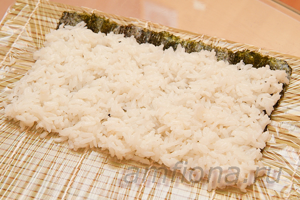 Распределите рис для суши, смачивая ладонь водой. Оставьте свободной небольшую полосу на ближнем к вам крае нори и сделайте смещение на дальнем. Прижимать рис не нужно, рисинки должны быть просто склеенными, а не спрессованными. 