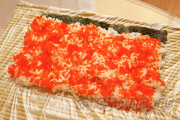 Пару столовых ложек оранжевой икры летучей рыбы распределите по всей поверхности риса. Накройте рис ковриком и переверните так, чтобы рис с икрой оказался внизу, а нори - сверху. 