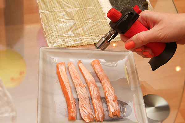 Нарежьте сёмгу для суши ломтиками. Если у вас есть газовая горелка, то можно обжечь пламенем рыбу, чтобы она осталась сырой внутри, но приобрела копчёный аромат.