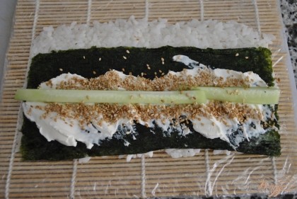 На циновке разложить мокрыми руками рис(2-3 стол.ложки). Сверху положить лист нори, намазать сыром, посыпать кунжутом, выложить огурец.