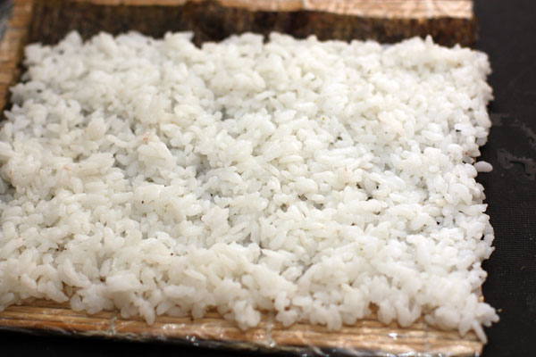 Половинку листа нори кладем на обернутый пищевой пленкой макису (коврик для суши). Сверху влажными рукми или ложкой выкладываем слой риса. При этом с дальней стороны оставляем 1 см нори свободным, а с ближней наоборот, рис должен заходить за нори на 1 см.