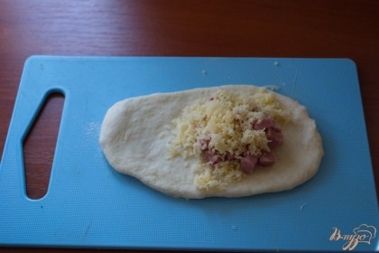 Кусочек теста раскатайте в лепешку. На край лепешки выложите предварительно нарезанные ветчину и натертый сыр.