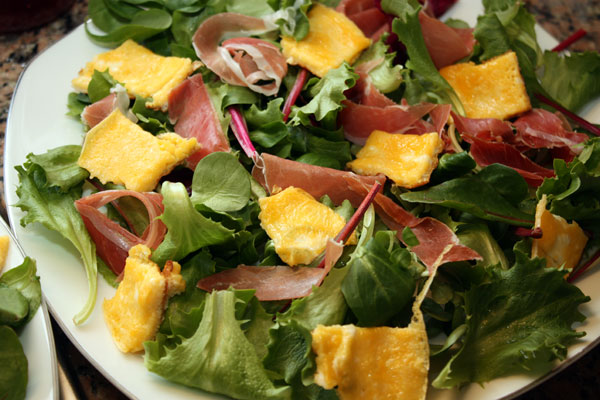 На широкую тарелку выложите салатные листья. Можно брать любые, которые вам по вкусу, лучше даже смесь. Важно, чтобы они были хрустящими и сочными.  Сверху разложите кусочки омлета и хамона.